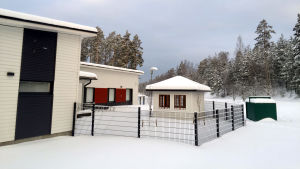 Byggnaderna i Gammelboda. Vit byggnad med mörgrå detaljer intill en inhägnad. Innaför staketet ett vitt litet hus. I bakgrunden skog.