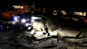 Arbetena vid järnvägsbron i Karis centrum är på målrakan. Bilden är från 8.10. Den anknyter med andra ord inte till kopparkablarna som gick av 9.10. En grävmaskin med stark ledbelysning jobbar i mörkret i ett centrumområde, sand och grus syns och man kan se ljus från skyltfönster.