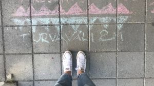Utanför dörren till allaktivitetshuset har något ritat var man ska stå för att hålla ett säkert avstånd till den som köar framför en. Någon har skrivit "turvaväli" med kritor på gatustenarna.