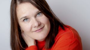 Marie Aubert är kommunikatör, journalist och författare och bosatt i Oslo. Porträttbild av Marie Aubert som tittar rakt in i kameran, iklädd en orange tröja.