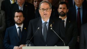 Quim Torra fördömer våldsamheterna i Barcelona. Kataloniens regionpresident Quim Torra håller tal. I bakgrunden ses ett antal män i mörka kostymer, likt den som Quim Torra bär.