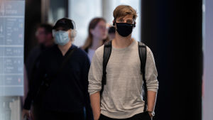 Vissa finländare har redan använt ansiktsskydd trots att det inte funnits en klar rekommendation om det. Mies kävelee kohti kameraa kasvoillaan musta suun ja nenän peittävä hengityssuojain. Miehen takana kävelee nainen lippalakki päässä jolla on myös hengityssuojain. Taaempana kävelee kohti muita ihmisiä, joilla ei ole hengityssuojamia.
