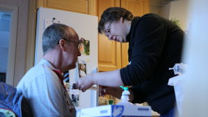 Anna Wadén ger Tomas Groop antibiotika genom en centralvenkateter. En man sitter hemma i sitt kök och får medicin genom en kateter av en sjukskötare.