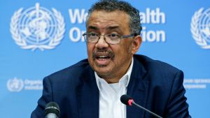 WHO:s generalsekreterare Tedros Adhanom Ghebreyesus tal avbröts av en hostattack, men han försäkrade att det inte var frågan om corona En man i glasögon och kostym sitter bakom ett par mikrofoner med WHO:s logga i bakgrunden.