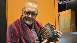 Johan Fröberg. Johan Fröberg, en man med glasögon och rödblårandig morgonrock, lyssnar på en gammal radio han har i famnen.