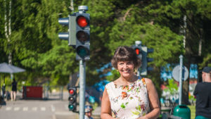 Minister Berner vill se mer äldre elever hjälpa de yngre i trafiken. En glad kvinna står på en asfalterad gångbana. I bakgrunden syns ett barn på en cykel och ett rött trafikljus.