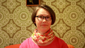 Minna Sarantola-Weiss är expert på soffgruppen i finska hem. Kvinna i brun page och glasögon mot en vägg med mönstrad, grön tapet i 70-talsstil. Fotad på Helsingfors stadsmuseum.