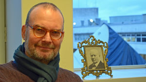 Museichef Kai Kartio. Porträtt på museichef Kai Kartio med foto på Amos Andersson i bakgrunden.