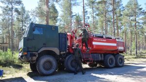 Den här militära brandbilen kan ta sig ut i skogen i Syndalen för att släcka bränder. En bild på två beväringar som klättrar upp på en tankbil som fungerar som brandbil. Tankbilen är röd och det står Puolustusvoimat (Försvarsmakten) på den. Runt omkring den finns det tallskog.