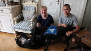 Kia och Alexander Brummer med hundarna Nalle och Astro. En kvinna och en ung man sitter på golvet och leker med två stora svarta diabeteshundar.