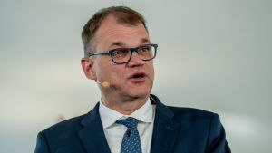 Juha Sipilä Juha Sipilä talar i en mikrofon, en såkallad mygga, på kinden. Han har kostym och glasögon.