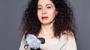 Nahid Persson är både framför och bakom kameran i filmen. Dokumentaristen Nahid Persson mot grå vägg med videokamera på stativ.