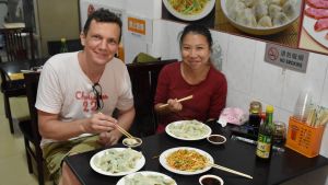 Tuomas Harjumaaskola och hans hustru Miao har med egna ögon kunnat följa med hur snabbt antalet elbilar har ökat. I Shenzhen är de allra vanligast. Fotografen Harjumaaskola poserar tillsammans med en kvinna framför ett matbord fyllt med kinesiska dumplings