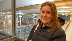 Alli Liljeström kommer säkert besöka simhallen under sportlovet också. En kvinna med ytterkläder och vått hår står inomhus och tittar in i kameran. Bakom henne syns ett fönster och man ser ner mot en simbassäng.