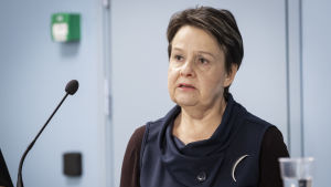 Social- och hälsovårdsministeriets kanslichef Kirsi Varhila. En kvinna i medelåldern med kort mörkt hår och mörka kläder ser allvarlig ut och ser ut att prata där hon står framför en mikrofon och tittar snett ut ur bilden.