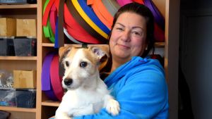 Jaana Alén med sin Jack russell terrier Milou. En kvinna står med en liten, ljus hund i famnen, i bakgrunden syns rullar med remmar i olika färger.