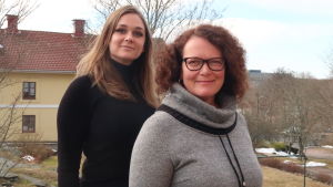 Alexandra Lönnström (till vänster). En svartklädd ung kvinna med långt brunt hår står bakom en medelålders kvinna i mörkt hår med korkskruvade hårlockar, glasögon med svarta bågar och grå tröja. De står framför ett gult stenhus i tidigt vårväder.