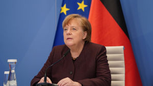 Förbundskansler Angela Merkels strikta linje har väckt kritik hos oppositionen. En kvinna i rödaktig jacka sitter med en mikrofon framför sig. Hon har sina händer på bordet. Bakom henne syns två flaggor.