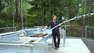 Joacim Mäkelä demonstrerar hur hårt vattentrycket är. Man klädd i svart vid modern mattvätt på betongyta. I bakgrunden skog. Mannen sprutar vatten ur en av slangarna vid tvätten.
