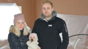 En familj bestående av kvinna, man och en liten baby står i vinterkläder inne i ett halvfärdigt nytt hus.