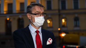 Justitiekansler Tuomas Pöysti konstaterar att beredskapslagen är en sista utväg. Tuomas Pöysti hämärtyvässä illassa Senaatintorilla. Tuomas on pukeutunut tummansiniseen pukuun. Vaaleansinisen kauluspaudan parina on punainanen kravatti ja rintataskusta pilkistää punaisilla ympyröillä koristeltu valkonen liina. Kasvomaski on käytössä.