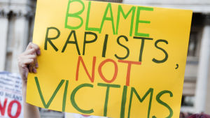 Nainen pitää mielenosoituksessa ylhäällä kylttiä jossa lukee: Syyttäkää raiskaajaa, älkää uhria 
