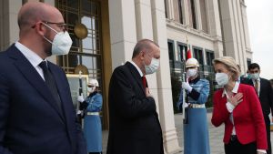Turkiets president Recep Tayyip Erdoğan (i mitten) tog emot EU-kommissionens ordförande Ursula von der Leyen och Europeiska rådets ordförande Charles Michel i Ankara. Två kostymklädda män och en kvinna i röd jacka och svarta byxor står utanför en byggnad med två soldater med gevär och långa ljusblå vapenrockar bakom sig. Alla bär munskydd. Från vänster Charles Michel, Recep Tayyip Erdogan och Ursula von der Leyen.