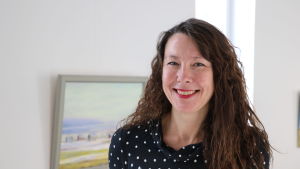 Ann-Sofi Backgren. En kvinna med långt brunt hår står framför en vit vägg. På väggen hänger en tavla.