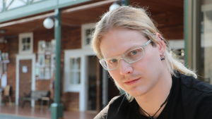 Rasmus Anderssén föreläser om självmord och hur de kan förebyggas. Han har själv överlevt ett självmordsförsök och vet att det finns hjälp att få. En ung man med långt ljust hår och glasögon sitter vid ett bord.