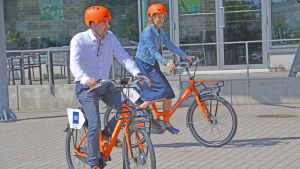 Trots de fina hjälmarna på bilden så måste man ha med sig en egen hjälm då man använder en lånecykel. En kvinna och man cyklar på orange cyklar med matchande hjälmar.