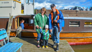 Anne och Per-Olof Johansson tog med sig barnbarnet Ian Ebeling till butiksbåten för första gången. En dam, en man och ett litet barn står på en brygga utanför en större motorbåt.