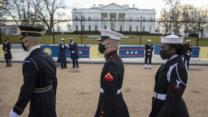 Militären övar inför dagens parad. Tre personer i USA:s armétruppers paraduniform marcherar förbi kameran. I bakgrunden syns vita huset. Det är två vita män och en svart kvinna på bilden.