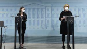 Marin och Haatainen på presskonferensen. Statsminister Sanna Marin (till vänster) och arbetsminister Tuula Haatainen vid regeringens presskonferens. Båda har munskydd på sig och de står vid varsitt talarpodium.