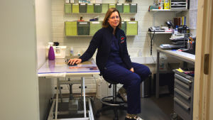 Veterinär Marina Hultholm Veterinär Marina Hulthom sitter på sitt rum på kliniken. Rummet är ganska litet och har ett undersökningsbord och ett skrivbord. Väggarna är vita.