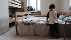 I flyktingförläggningar väntar fortfarande tusentals och åter tusentals asylsökande på besked om sin framtid. Många av dem är barn, som gjort de tunga resorna från Turkiet till Grekland i läckande gummibåtar. Pojke står vid säng och leker.