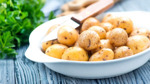 En vit skål med potatis och en liten kniv står på ett träbord med en knippe örter i bakgrunden.