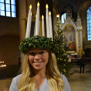 Åbos lucia 2020 och 2021, Janina Karrento, en dam med långt ljust hår och en ljuskrona på huvudet, ler mot kameran. Hon står i Åbo Domkyrka med en stor julgran i bakgrunden.