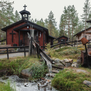Kolme pientä mökkiä, joista yhdellä on katolla risti. Mökkien editse virtaa puro.