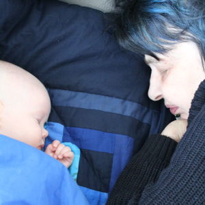 Iidan äiti nukkuu lapsenlapsensa vierellä. Hänellä on siniset hiukset ja musta villapaita.