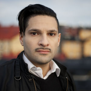 Karim Jebari vid Institutet för framtidsstudier porträtt av karim jebari. han bär en mörk jacka med en vit skjortkrage som sticker upp under jackan. karim bär en tinn mustasch och har mörkt hår. bilden är tagen utomhus i kvällsljus.