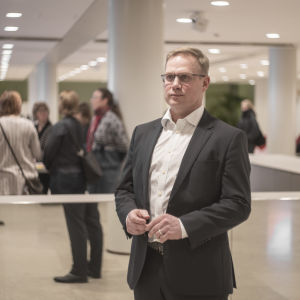 Niclas Grönholm ser optimistiskt på vårens rekryteringar, och hoppas att Helsingfors kommer att hitta den dagispersonal som behövs. En man står framför en spegel och några andra människor. Han ser till höger. Han har på sig en kostym och ser allvarlig ut.