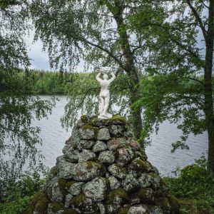 Valkoinen pieni miespatsas selin kameraan kiviröykkiön päällä, taustalla järvimaisemaa