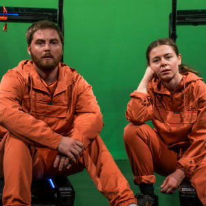 Skådespelarna Ole Øwre och Elisa Makarevitch sitter på marken iklädda orange overaller, i bakgrunden syns en grön skärm.