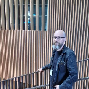 För varje generation är det lite olika saker som är viktiga och de hör till de äldre generationerna att vara kritiska till vad de yngre tycker att är viktigt, säger Rasmus Vuori. Rasmus Vuori står mörkklädd och håller i en ledstång mot en vägg av spjälverk i trä i aulan till Aalto-universitetet.