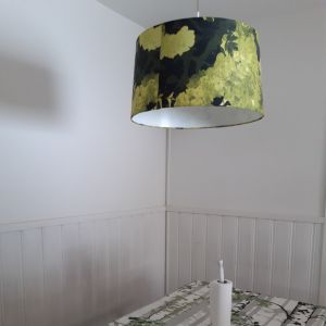Här kring köksbordet börjar skolmorgonen. Köksbord med duk i grönt och vitt som föreställer björkstammar, vit panelad vägg bakom köksbordet och en taklampa med grön skärm.
