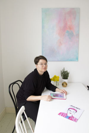 Tavlan bakom Zarah har gjorts under konstterapin. Zarah Holmberg sitter vid ett bord och ritar med sin ipad. På bordet ligger också tidningen hon gjort.