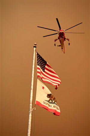 En helikopterlyfter mot föra att ge siv av mot brandbekämningsuppdrag. I förgrunden USA:s flagga och den kaliforniska delstatsflaggan.