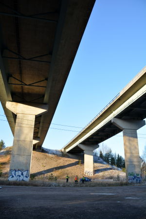 Personbilen körde av motorvägen och störtade ner under bron. Räddningspersonal och polis vid demolerad bil under motorvägsbro.
