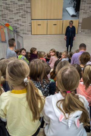 Rektor Carola Wiksten håller samling för ettorna. Hon berättar bland annat att skolan har kostat lika mycket som åtminstone 40 vanliga hus, och att det är viktigt att inte klottra eller förstöra saker i skolan. Morgonsamling, i förgrunden elever som sitter med ryggarna vända mot kameran i en trappa, rektorn står längst ner