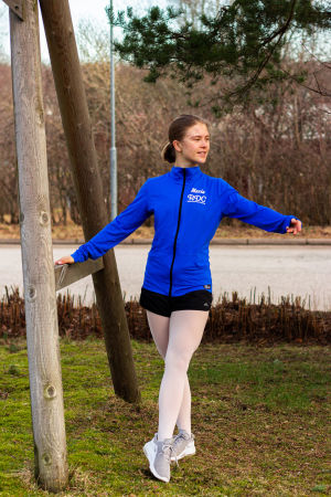 Maria Knaapinen håller en kort paus från datorn och skoluppgifterna. Hon passar på att sträcka på benen och göra en tendu, det vill säga ett danssteg inom baletten. En ung kvinna i blå sportjacka och vita trikåer gör en balettrörelse (tendu) utomhus. Tar stöd av en stolpe
